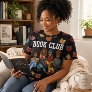 May include: Black t-shirt with the words "BOOK CLUB" in white. The shirt features illustrations of women reading books and scattered book graphics. The person is holding a book and sitting in a chair.
