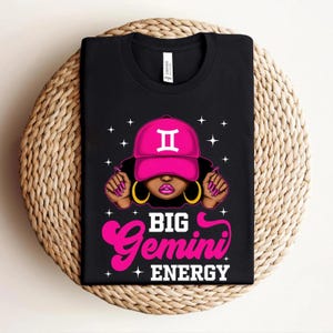 May include: Black t-shirt with a pink Gemini hat graphic and gold earrings. The text "BIG Gemini ENERGY" is in pink and white. The shirt is displayed on a woven surface. The design is a bold statement.
