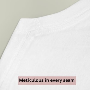 May include: Close-up of a white t-shirt neckline and shoulder. The text "Meticulous in every seam" is displayed on a pink rectangular label. The image highlights the garment's construction and attention to detail.