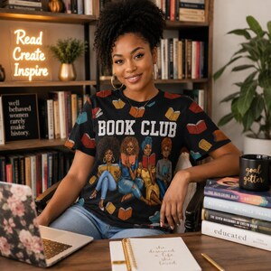 May include: A woman wearing a black t-shirt with the words "BOOK CLUB" and an illustration of women reading. The shirt is surrounded by books and a neon sign that says "Read Create Inspire". A black mug with the words "goal getter" is also visible.
