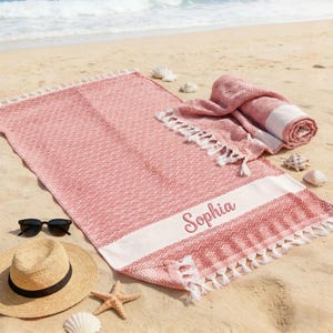 May include: A coral-colored beach towel with a white stripe and the name "Sophia" embroidered in pink. The towel has a geometric pattern and white fringe. A rolled-up towel, sunglasses, a straw hat, and seashells are on the sand.