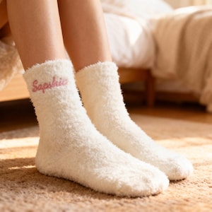 May include: A pair of cream-coloured fluffy socks. The word "Saphire" is embroidered in pink on the cuff of each sock. The socks are worn on a person's feet, resting on a light brown carpet.