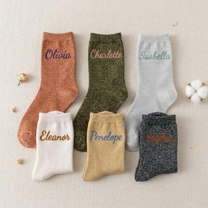 Peut inclure: Six paires de chaussettes scintillantes de différentes couleurs, chacune brodée d'un nom : Olivia, Charlotte, Isabella, Eleanor, Penelope et Sophia. Les chaussettes sont de couleurs orange, vert, argent, blanc, or et noir.