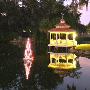 May include: A nighttime scene features a gazebo and a Christmas tree-shaped light display reflected in a body of water. The gazebo is illuminated with warm lights, and the tree is outlined with red and white lights. Trees surround the water.