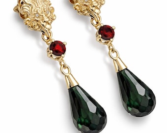 Peut inclure: Une paire de boucles d'oreilles pendantes dorées. Chaque boucle d'oreille présente un dessus orné et détaillé avec de petites pierres claires, une pierre rouge et une pierre en forme de goutte d'eau vert foncé facettée. Les boucles d'oreilles sont présentées sur fond blanc.