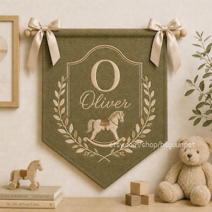 May include: Olive green wall hanging embroidered with the name "Oliver" in cream thread. Features a rocking horse and laurel wreath design. Includes cream bows and wooden dowels for hanging. A teddy bear and wooden blocks are in the background.