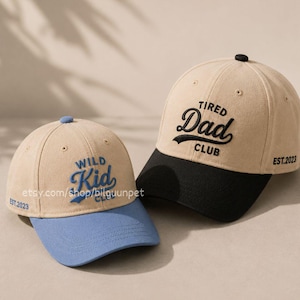 Pode incluir: Dois bonés de beisebol. Um é bege com aba preta e as palavras "Tired Dad Club" bordadas em preto. O outro é bege com aba azul e as palavras "Wild Kid" bordadas em azul. Ambos têm "EST.2023" na lateral.