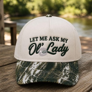 Puede incluir: Gorra de béisbol beige con el texto bordado "LET ME ASK MY Ol' Lady" en verde oscuro. La visera presenta un estampado de camuflaje en tonos verdes y blancos. La gorra está sobre una superficie de madera.