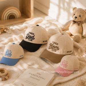 Pode incluir: Uma coleção de bonés de beisebol bege com abas contrastantes. Os bonés apresentam as frases "Tired Dad Club", "Tired Mom Club" e "Wild Kid Club". Cada boné tem o ano "EST.2023" bordado. Um urso de pelúcia e um livro também estão na imagem.