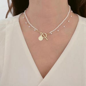 Op de afbeelding: Een gelaagde ketting met kleine, witte parels en een goudkleurige ketting. De ketting heeft een tuimelsluiting en een kleine, ronde hanger met een gedetailleerd ontwerp. De ketting wordt gedragen op een witte top.