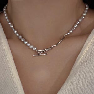 May include: A silver necklace with a strand of grey pearls. The necklace has a distinctive design, with a wavy silver chain and a T-bar clasp. The pearls are irregularly shaped, enhancing the necklace's organic aesthetic.