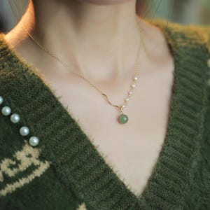 Könnte beinhalten: Eine zarte goldene Halskette mit einem kleinen, runden, grünen Jade-Anhänger. Die Halskette hat ein wellenförmiges Design und ist mit kleinen Perlen verziert. Die Person trägt einen grünen Pullover.