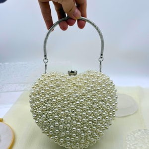 May include: A heart-shaped handbag covered in off-white pearls. The bag has a silver-toned handle and clasp. The pearls are of varying sizes, creating a textured surface. The bag is a romantic accessory.