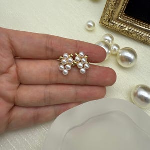 May include: Gold-coloured stud earrings, each with a cluster of small, round, white pearls. The earrings are held in a hand, with other pearls and a gold-framed object in the background. The earrings have a floral design.