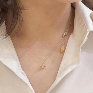 May include: A delicate gold necklace featuring a small planet pendant with a pearl and a dangling oval charm. The necklace is worn against a white collared shirt.