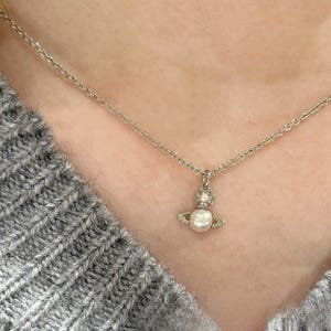 Puede incluir: Un collar plateado con un pequeño colgante. El colgante presenta una perla y un diseño de cruz. El collar se lleva sobre un jersey gris. El colgante tiene forma de planeta con una perla en el centro.