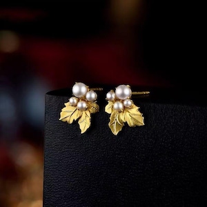 May include: A pair of gold-colored stud earrings. Each earring features a cluster of white pearls atop a gold leaf design. The earrings are displayed on a black surface against a blurred, dark background.