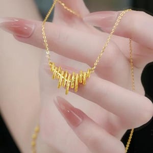 May include: A gold-colored necklace with a unique design. The pendant features a series of vertical bars arranged in a curved shape. The chain is delicate and complements the pendant. The necklace is likely made of gold or a gold-toned metal.
