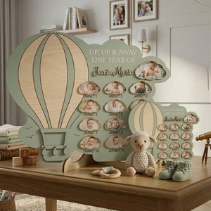 Puede incluir: Un expositor de fotos de madera con forma de globo aerostático, con el texto "UP, UP & AWAY ONE YEAR OF Santo Maria". Cuenta con espacios para fotos y está decorado con un pequeño cordero de peluche y zapatos de bebé. El expositor está sobre una mesa de madera.
