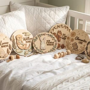 Puede incluir: Placas de nombre de madera para guardería con diseños de animales grabados, incluyendo un zorro, una nutria, un conejo, una ardilla, un ratón y un ciervo. Cada placa presenta un nombre. Las placas se exhiben en una cuna blanca.