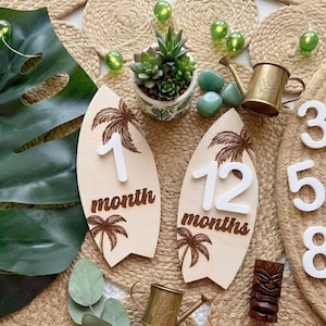 Könnte beinhalten: Meilenstein-Marker in Form von Surfbrettern aus Holz mit den Zahlen 1 und 12 sowie den Wörtern "month" und "months" in brauner Schrift. Die Bretter haben ein Palmenmuster. Weiße Zahlen 3, 4, 5, 6, 7 und 8 sind ebenfalls vorhanden.