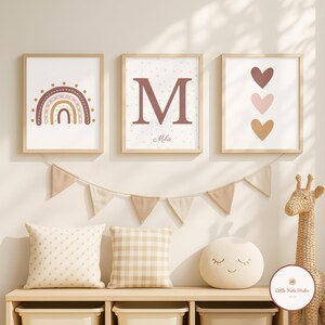 Peut inclure: Trois impressions encadrées avec une palette de couleurs neutres. L'une présente un arc-en-ciel, une autre une grande lettre "M" avec le nom "Mila", et la troisième a trois cœurs. Une guirlande, des coussins et un jouet girafe complètent le décor de la chambre d'enfant.