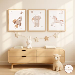 Peut inclure: Trois impressions d'art encadrées sur le thème de l'espace. Les impressions présentent une fusée, un ours en combinaison spatiale et une licorne. La palette de couleurs comprend des tons beige, marron et pêche. Une commode en bois, une guirlande d'étoiles et des animaux en peluche sont au premier plan.