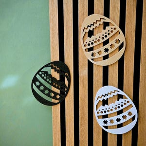 May include: Three decorative Easter egg cutouts in black, beige, and white, each with unique geometric patterns. The eggs are displayed against a wooden slatted background and a green wall.