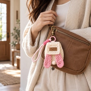 Puede incluir: Un llavero de mini mochila de ganchillo color crema con largas orejas de conejo rosas, una pequeña cola de pompón rosa y un parche rectangular de aspecto de cuero marrón en la solapa frontal, sujeto a un bolso bandolera marrón con un cierre metálico.