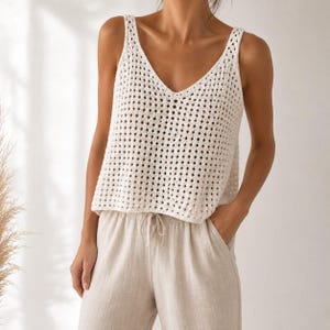 May include: A sleeveless white crochet tank top featuring a V-neckline and an open diamond-shaped knit pattern. The top is paired with light beige drawstring trousers that have a relaxed fit and a visible tie at the center of the waistband.