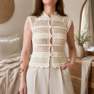 May include: A sleeveless, cream-colored crochet top featuring a delicate openwork pattern, a small collar, and a front button-down closure with five round buttons. The garment has a scalloped hemline and a fitted silhouette, crafted from yarn.