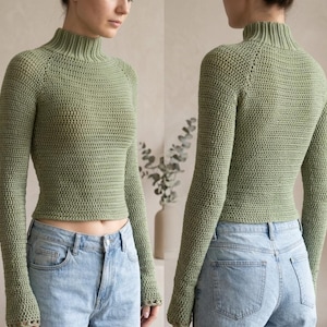 Puede incluir: Un suéter de manga larga en color verde salvia con cuello alto acanalado y un patrón de tejido de ganchillo texturizado. La prenda es ajustada, cuenta con mangas raglán y un dobladillo corto, combinada con jeans de mezclilla azul claro.
