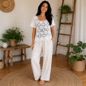 Può includere: Un top bianco a maniche corte caratterizzato da un intricato motivo floreale all'uncinetto con scollo e orlo smerlati, abbinato a pantaloni beige larghi e comodi. Il top ha una consistenza semi-trasparente, mentre i pantaloni sembrano realizzati in tessuto opaco.