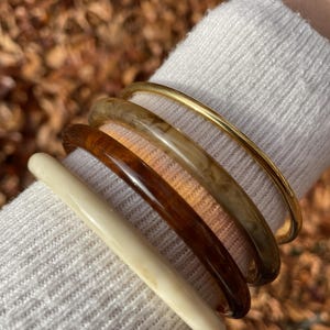 Könnte beinhalten: Eine Sammlung von Armreifen in verschiedenen Farben und Materialien. Das Set enthält einen goldfarbenen Metallarmreif sowie mehrere Harzarmreifen in Creme-, Braun- und Marmortönen. Die Armreifen sind auf einem weißen Pulloverärmel gestapelt.