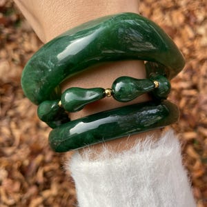 Puede incluir: Tres pulseras de jade de varios estilos. Las pulseras superior e inferior son anchas, lisas y de un verde esmeralda intenso. La pulsera central presenta cuentas en forma de lágrima con detalles dorados. Las pulseras se llevan en una muñeca con un suéter blanco y difuso.