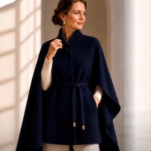 Op de afbeelding: Een donkerblauwe cape met een hoge kraag en een bijpassende strikceintuur in de taille. Het kledingstuk heeft wijde armopeningen en een gladde, effen stofconstructie. Het wordt gedragen over een crèmekleurige top met lange mouwen en ribbelstructuur.