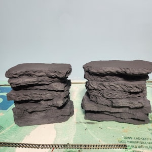 May include: Two stacks of gray, layered rock formations, likely for a model railroad or diorama. The rocks have a rough, textured surface and are arranged on a green and white surface with model train tracks.