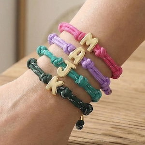 Peut inclure: Une main portant cinq bracelets en corde colorés. Chaque bracelet est d'une couleur différente : vert foncé, sarcelle, violet et rose. Chaque bracelet a un charme de lettre dorée : K, J, A et M. Les bracelets sont sur une surface en bois.