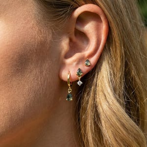 May include: A close-up of a person's ear adorned with three sets of earrings. The earrings include small studs, a pair with a dangling teardrop-shaped green gem, and a pair of gold hoop earrings with a teardrop-shaped green gem and a small diamond.