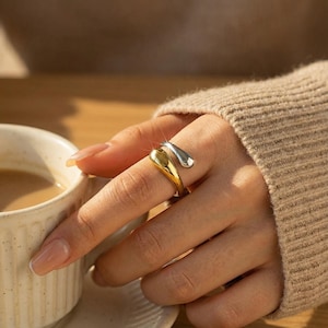 May include: A gold and silver-toned ring with a unique, flowing design. The ring features a gold base that wraps around the finger, with a silver-colored accent that curves over the top. The jewelry is worn on a hand, with a cup of coffee in the background.