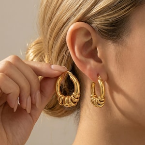 Puede incluir: Pendientes de aro dorados. Los pendientes tienen un diseño grueso y redondeado con una serie de pequeños detalles enrollados a lo largo de la mitad inferior del aro. Un pendiente es sostenido por una mano, mientras que el otro se usa en una oreja.