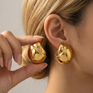 以下が含まれることがあります： 前面に斜めの溝が入った、金色のしずく型のイヤリング。イヤリングは光沢のある反射面を持っています。片方のイヤリングは手で持ち、もう片方は耳に装着されています。