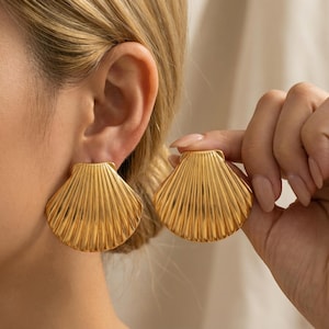 May include: Gold-colored seashell earrings. The earrings are large, with a detailed, ridged design. One earring is being held up by a hand, while the other is being worn on an ear. The earrings have a shiny, metallic finish.