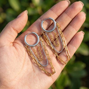 Puede incluir: Pendientes de aro plateados con cadenas colgantes doradas y plateadas. Los pendientes presentan una mezcla de estilos de cadena, creando un efecto de capas. Los aros se sostienen en una mano. Un accesorio elegante.