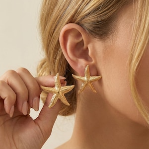 May include: Gold-colored starfish stud earrings. The earrings are shaped like starfish with textured surfaces. One earring is being held up by a hand, while the other is worn on an ear. The earrings are a beach-themed accessory.