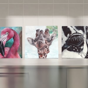 May include: Three framed animal paintings. The first painting features a pink flamingo with a green background. The second painting features a giraffe with a white background. The third painting features a black and white zebra with a close-up of its eye.