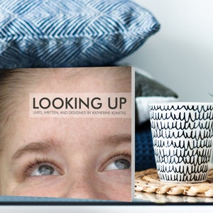 May include: A book cover with the title "LOOKING UP" and the text "Lived, written, and designed by Katherine Klumbitas". The cover features a close-up of a person's face looking up.