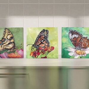 May include: Three framed watercolor paintings of butterflies on flowers. The butterflies are a swallowtail, a monarch, and a fritillary. The flowers are pink, red, and white.