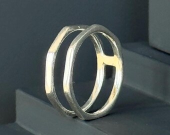 Könnte beinhalten: Ein silberfarbener Ring mit einem einzigartigen Doppelband-Design. Der Ring hat zwei parallele Bänder, die durch vertikale Stege verbunden sind, wodurch eine offene, geometrische Ästhetik entsteht. Der Ring wird auf einer dunklen Oberfläche präsentiert.