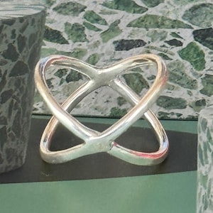 Può includere: Un anello color argento con un design intrecciato, che ricorda due cuori sovrapposti. L'anello è esposto su una superficie verde, con uno sfondo strutturato e due piedistalli cilindrici.